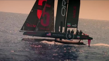 A high-speed racing catamaran sailing yacht with the number 5 and Oracle Team USA markings, cutting through open sea water. From the Top Gear New Zealand race where a rental car competes against the yacht.