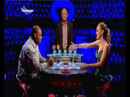 A still from the British TV game show Split or Steal, showing the host standing between two contestants at a round table with stacks of gold bars, illustrating a simultaneous move game where both parties must choose their actions without knowing the other's decision.